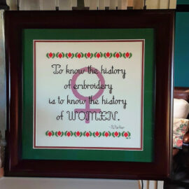 History Lesson: a framed cross stitch sampler. The words "To know the history of embroidery is to know the history of women" are stitched over a pinky-violet Venus symbol. From Grimalkin Crossing.