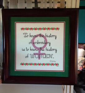 Herstory: a History Lesson in Cross Stitch: this is a framed cross stitch sampler. The words "To know the history of embroidery is to know the history of women" are stitched over a pinky-violet Venus symbol. From Grimalkin Crossing.