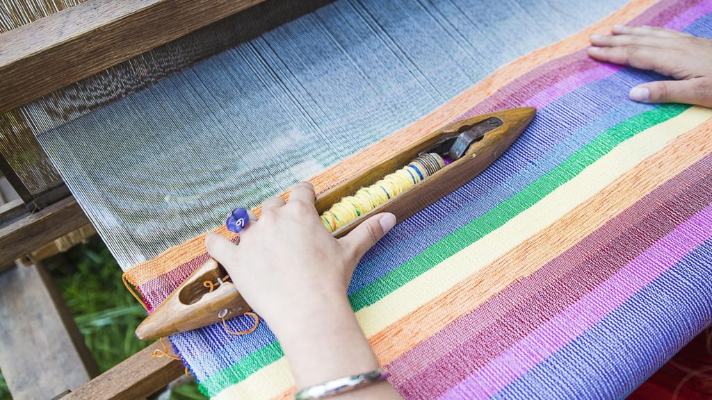 handmade decor. A pair of hands are weaving a rainbow on a traditional loom. From Grimalkin Crossing