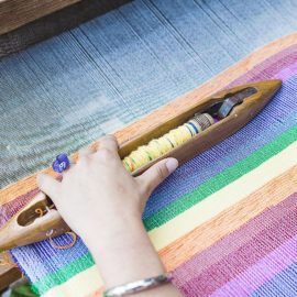 handmade decor. A pair of hands are weaving a rainbow on a traditional loom. From Grimalkin Crossing
