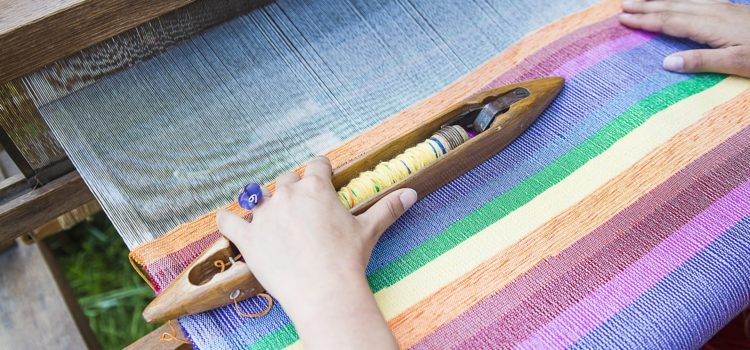 handmade decor. A pair of hands are weaving a rainbow on a traditional loom. From Grimalkin Crossing