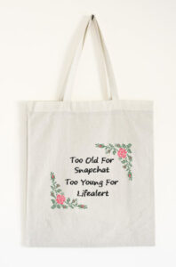 For Menopause Awareness Month. This image features a cross stitch sampler that's been stitched onto a tote bag. The legend "Too old for snapchat, too young for lifealert" is bracketed by a spray of ferns and roses on two corners. From Grimalkin Crossing.